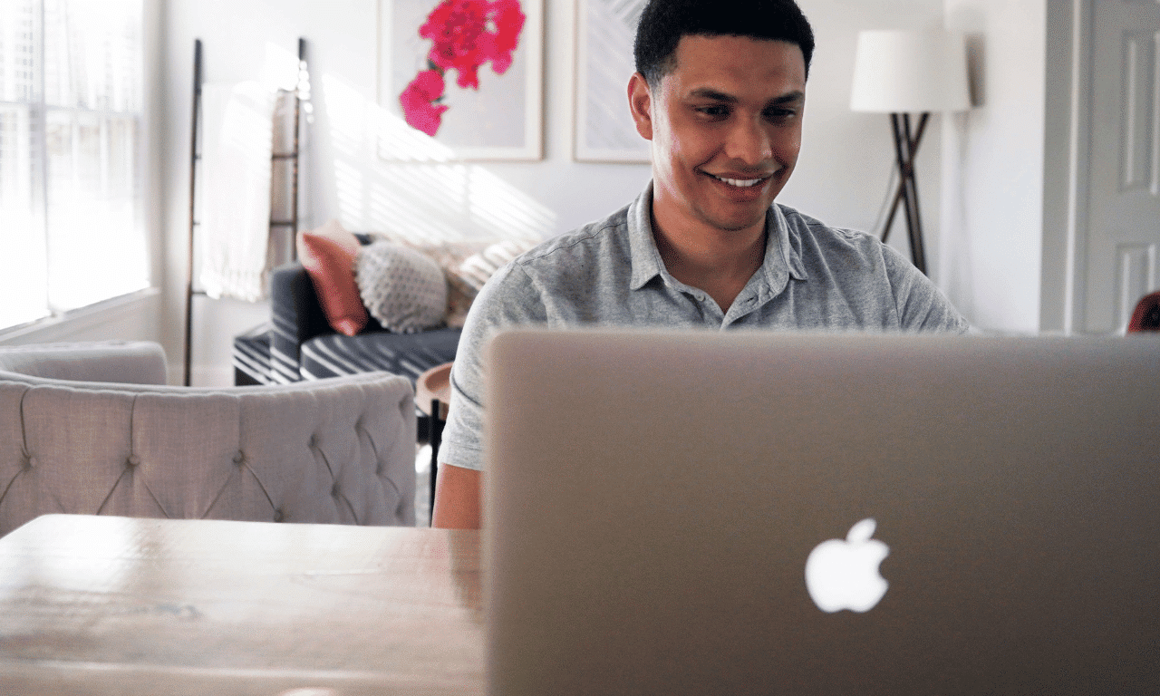 A male is sitting smiling in front of an Apple Macbook, in a light and airy living room. This represents Contractors.