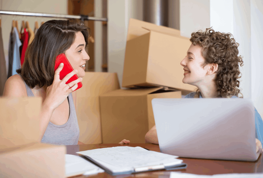 Two women smile at each other. One is holding a red phone to her ear, the other is sat in front of a laptop. A pile of boxes can be seen in the background. This represents online sellers.